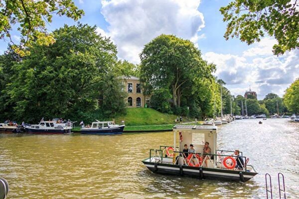 Menschen auf einer kleinen Fähre überqueren den Kanal in Leeuwarden, umgeben von Booten und üppigen grünen Bäumen, mit dem Oldehove-Turm im Hintergrund.